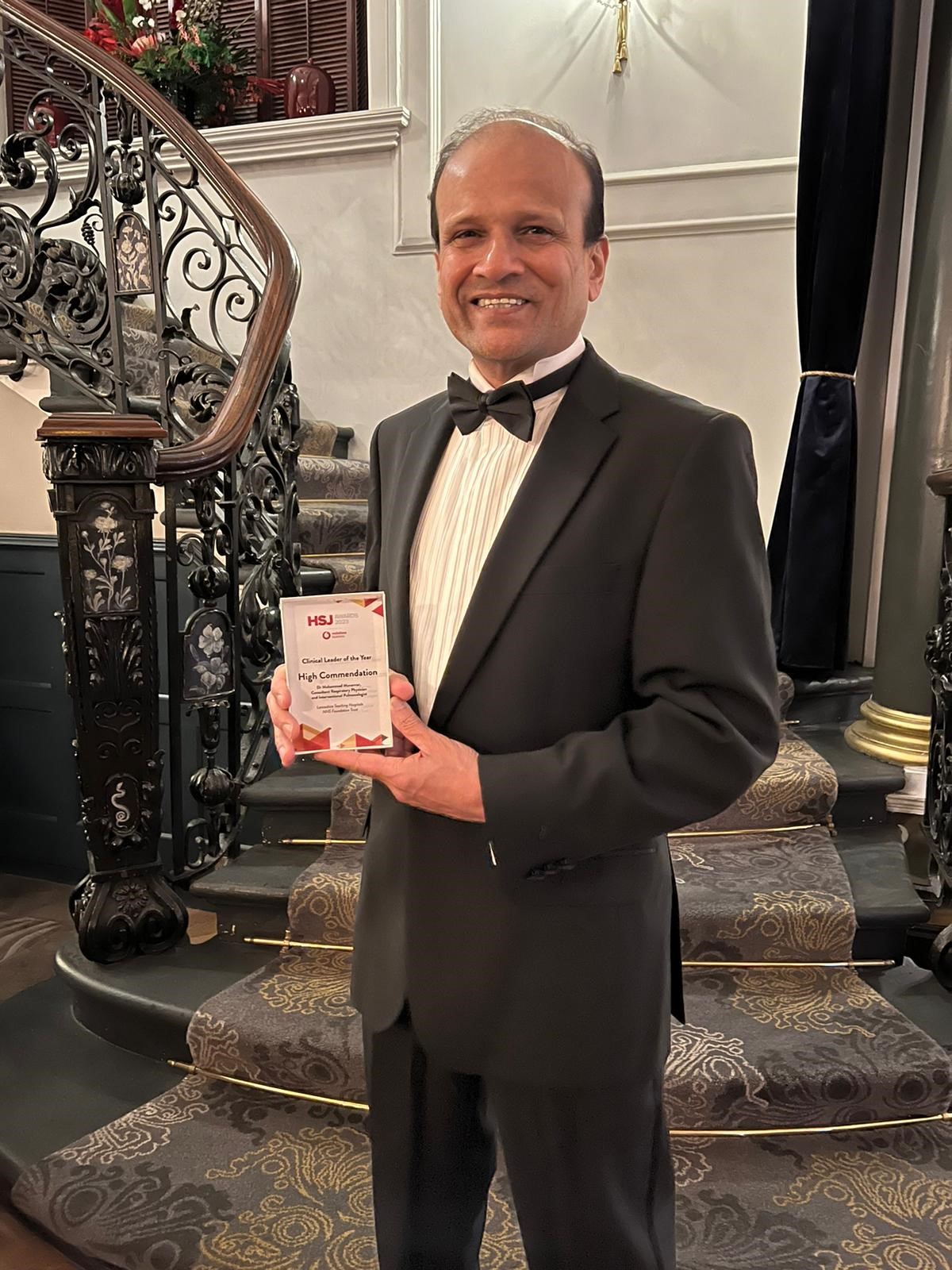 Dr Munavvar wearing a tux, smiling, holding an award plaque