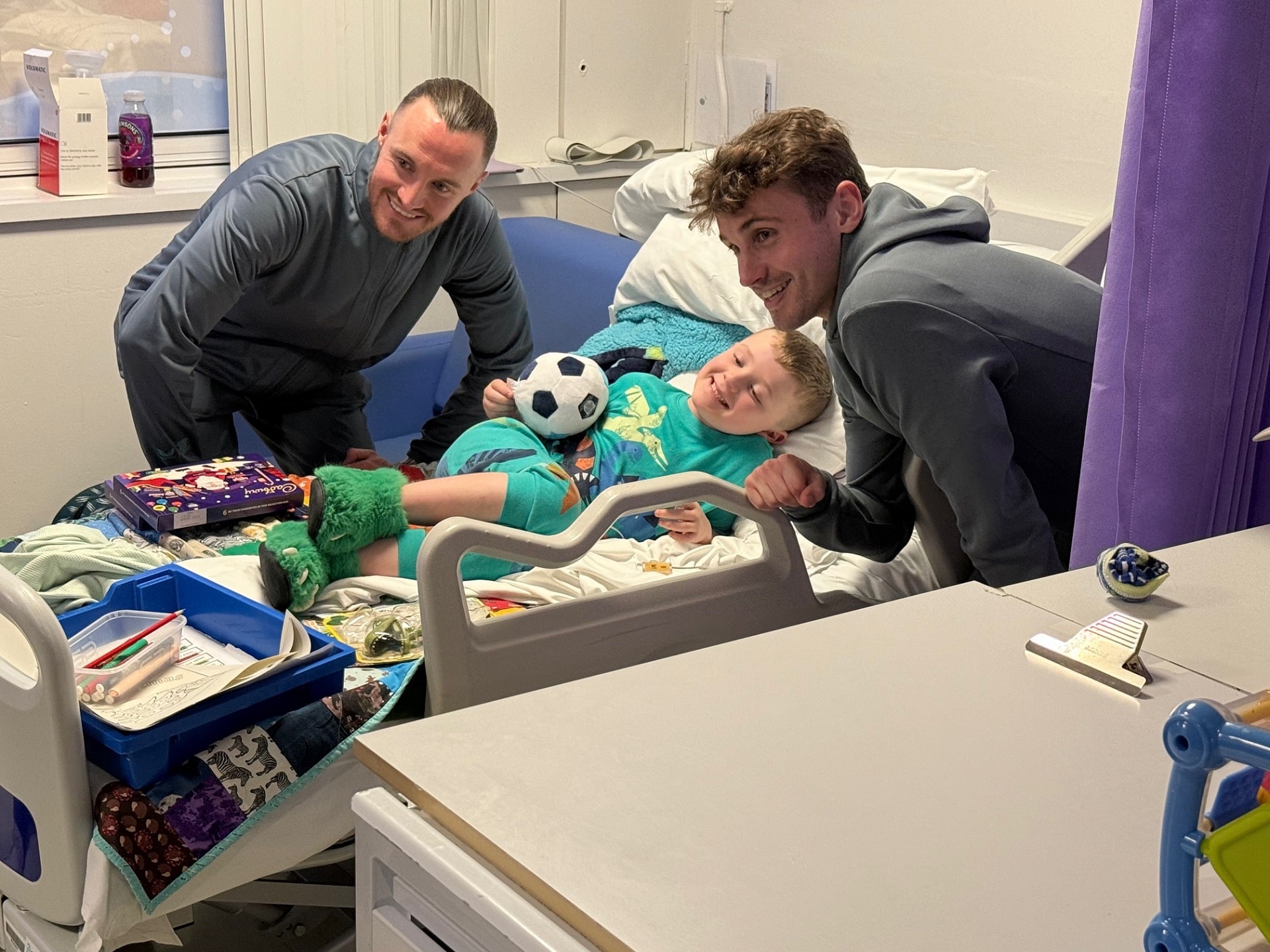 Photo of two PNE football players standing next to a smiling child on a medical bed