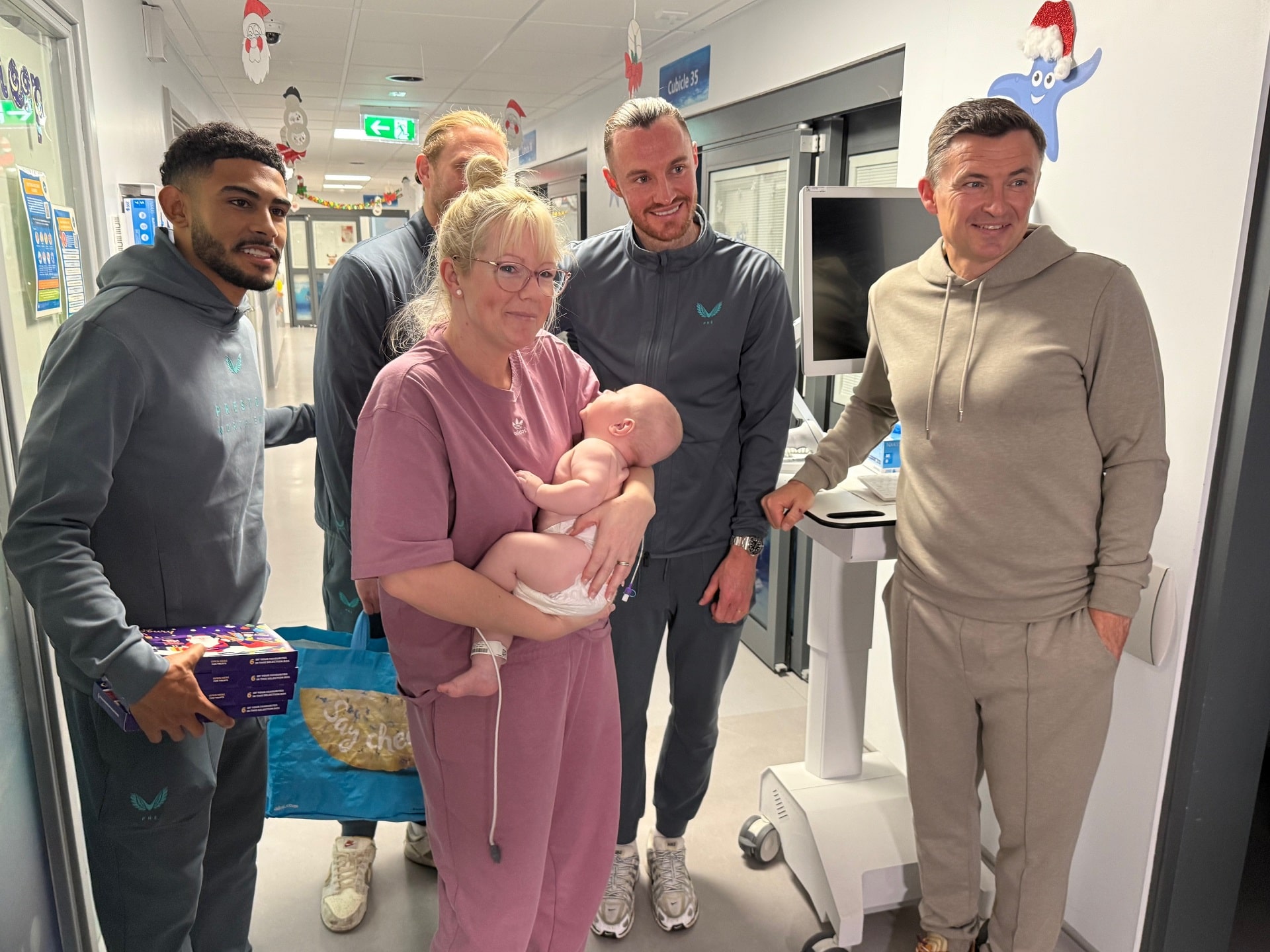 Photo of mother, holding her baby and smiling, surrounded by several PNE football players. All of them are in a hospital setting.