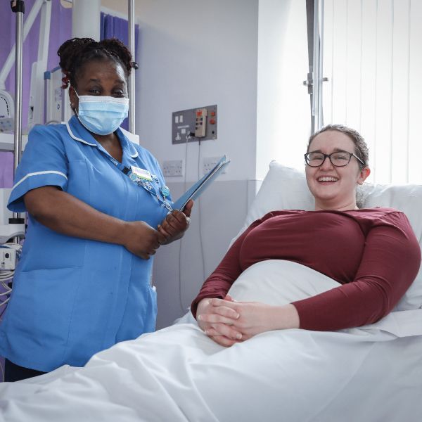 A photo of a uniformed NHS staff member standing next to a smiling patient in a hospital bed